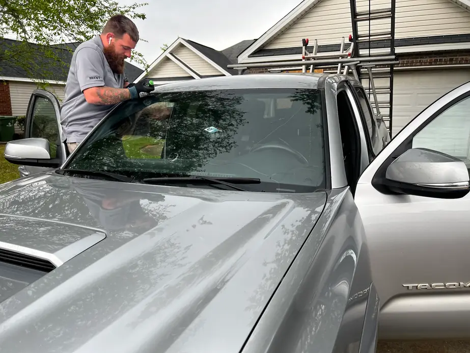 Brent in a grey work polo lifting a new windshield into a silver Toyota Tacoma on a driveway